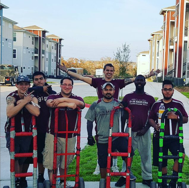 Six men in matching work shirts pose outdoors with hand trucks on a sidewalk, with apartment buildings and grass in the background. Six men in matching work shirts pose outdoors with hand trucks on a sidewalk, with apartment buildings and grass in the background.