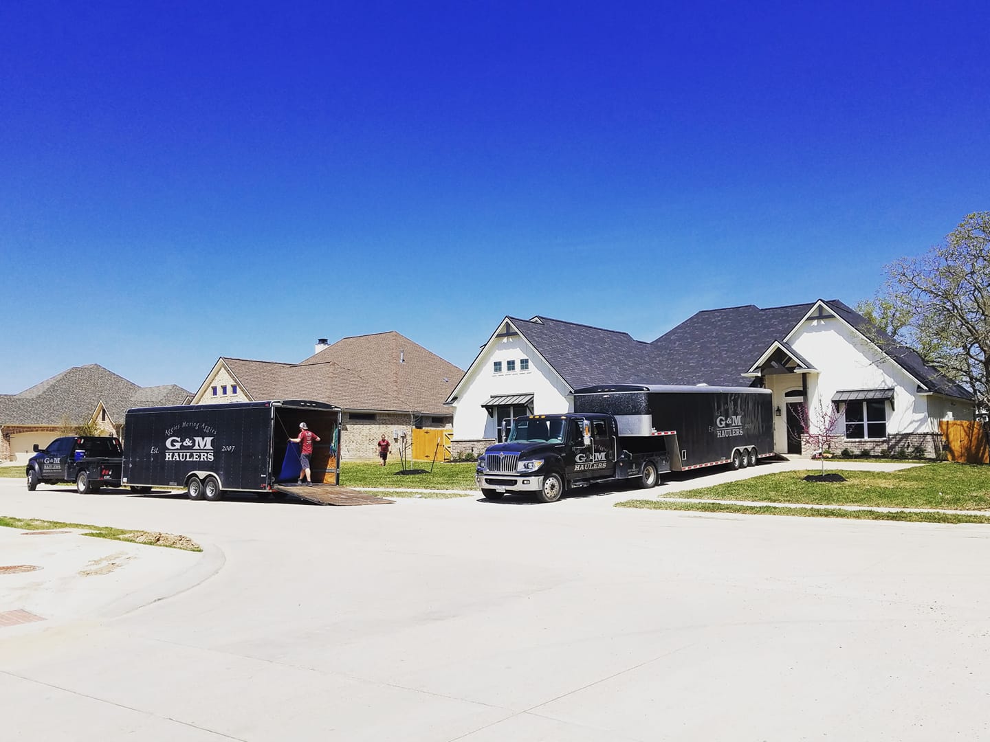 Two moving trucks and a trailer are parked outside a suburban house on a clear day. Movers are loading or unloading items near the vehicles. Two moving trucks and a trailer are parked outside a suburban house on a clear day. Movers are loading or unloading items near the vehicles.