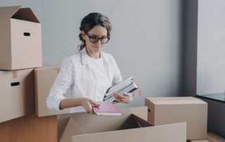 A woman wearing glasses packs books into a cardboard box, surrounded by other boxes in a room with natural light.